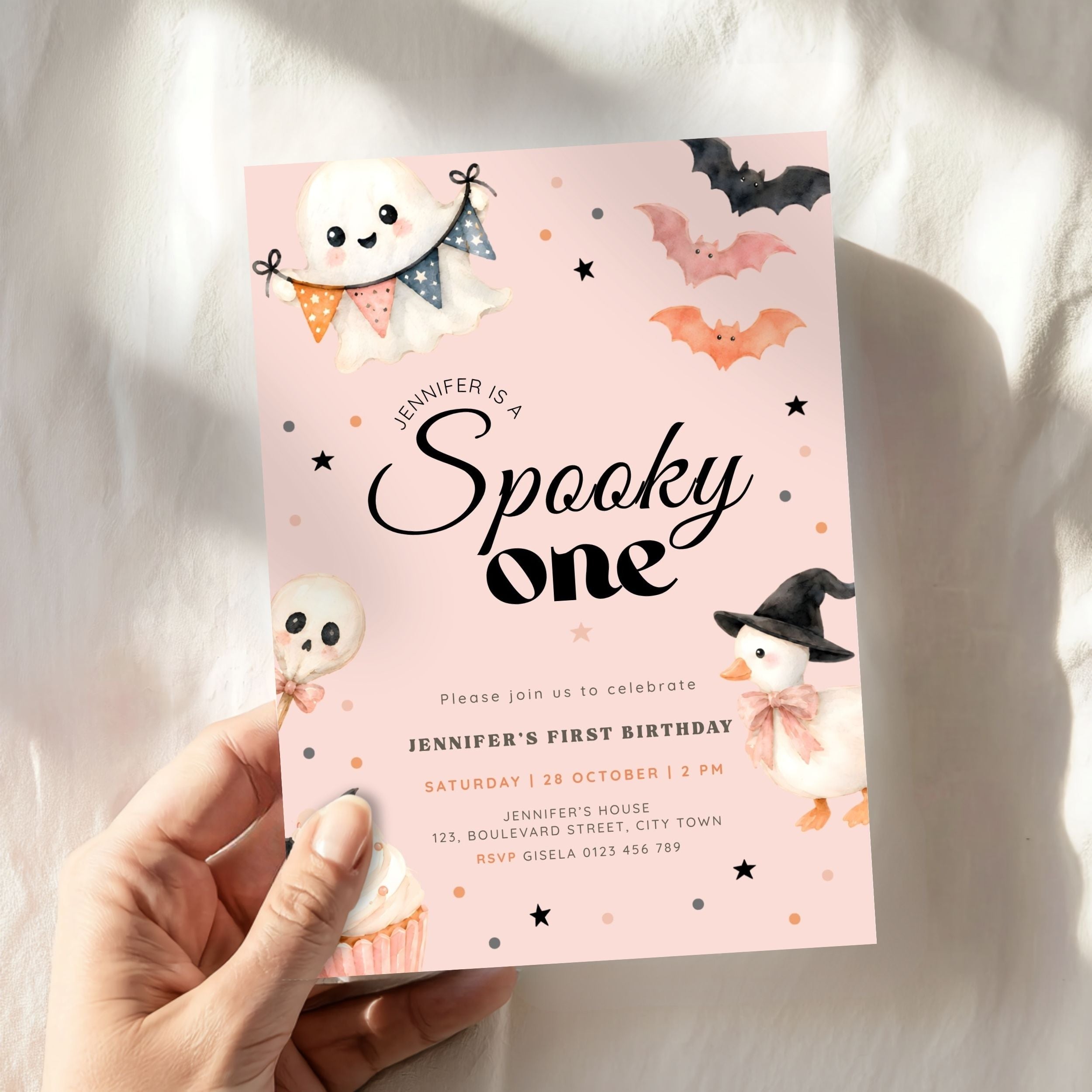A hand holds a pink Halloween first birthday invitation card featuring cute cartoon illustrations of a ghost, witch pumpkin, bat, and skull on a soft white fabric background.