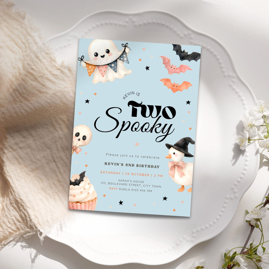 A Halloween-themed birthday invitation card styled on a white scalloped plate with soft cream fabric and white flowers around it. The card has a light blue background with illustrations of ghosts, bats, a penguin in a witch hat, stars, and a cupcake.