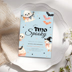 A Halloween-themed birthday invitation card styled on a white scalloped plate with soft cream fabric and white flowers around it. The card has a light blue background with illustrations of ghosts, bats, a penguin in a witch hat, stars, and a cupcake.