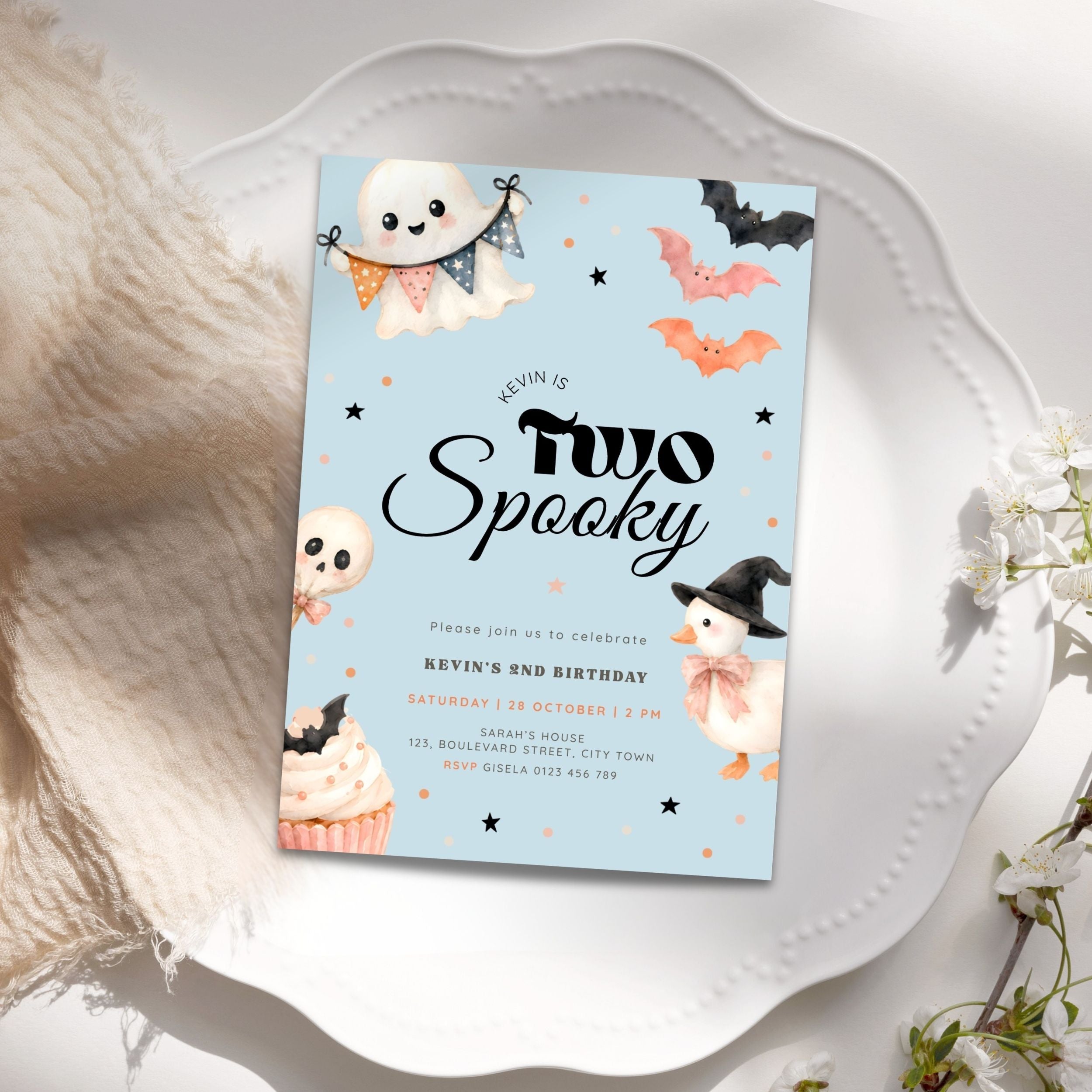 A Halloween-themed birthday invitation card styled on a white scalloped plate with soft cream fabric and white flowers around it. The card has a light blue background with illustrations of ghosts, bats, a penguin in a witch hat, stars, and a cupcake.
