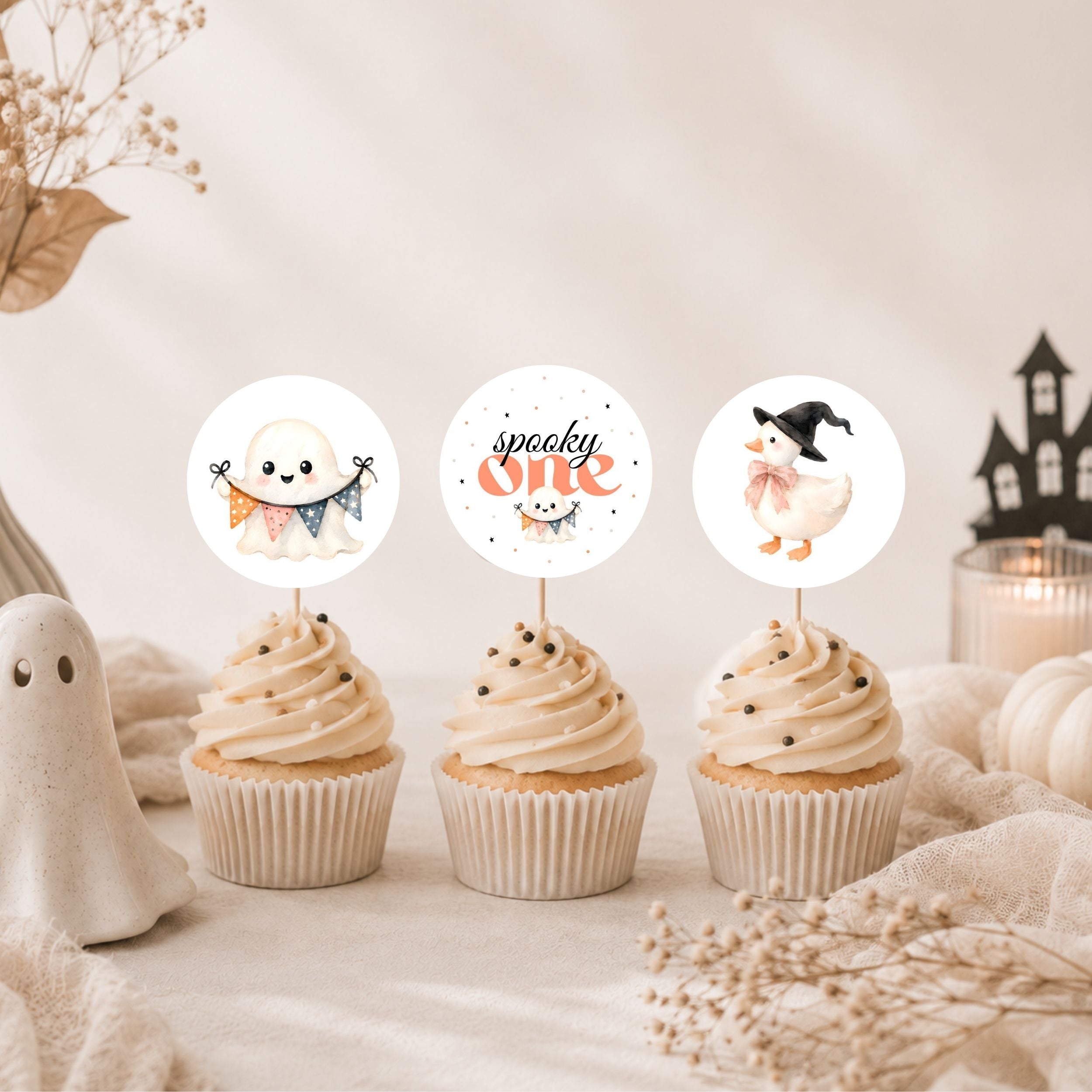 Three Halloween-themed cupcake toppers displayed on top of frosted cupcakes. The toppers feature cute illustrations: a smiling ghost with a banner, the words 'spooky one' with a pumpkin, and a duck wearing a witch hat.
