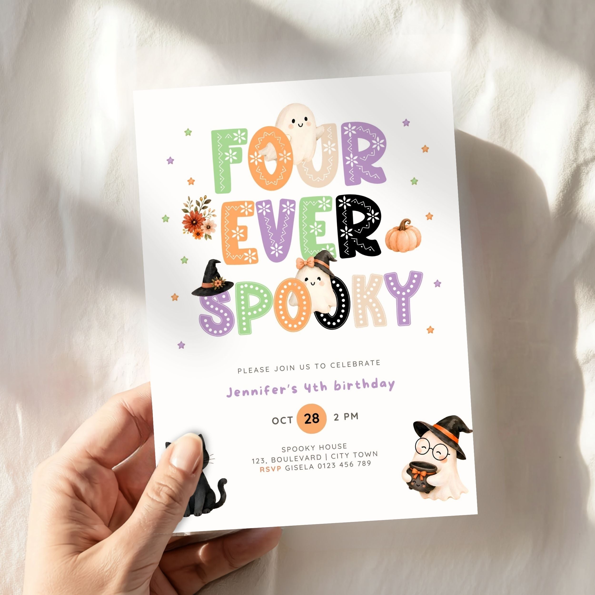 A hand holds a white Halloween-themed birthday invitation card with colorful playful lettering reading 'FOUR EVER SPOOKY' and cute cartoon illustrations, against a softly lit white fabric background.