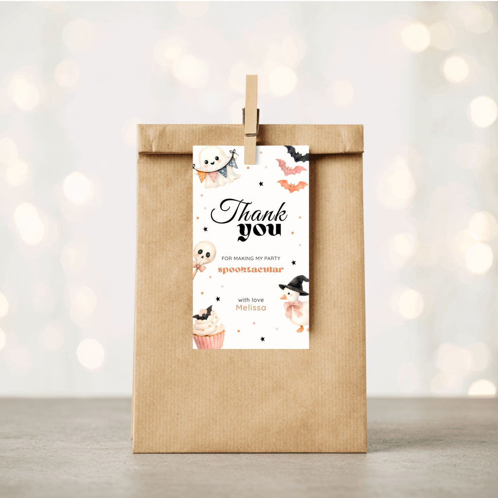 A kraft paper gift bag stands upright on a light surface with a wooden clip holding a printed thank-you tag. The tag has Halloween-themed illustrations and the text 'Thank you for making my party spooktacular' with the name 'Mia'.