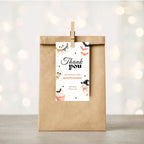 A kraft paper gift bag stands upright on a light surface with a wooden clip holding a printed thank-you tag. The tag has Halloween-themed illustrations and the text 'Thank you for making my party spooktacular' with the name 'Mia'.