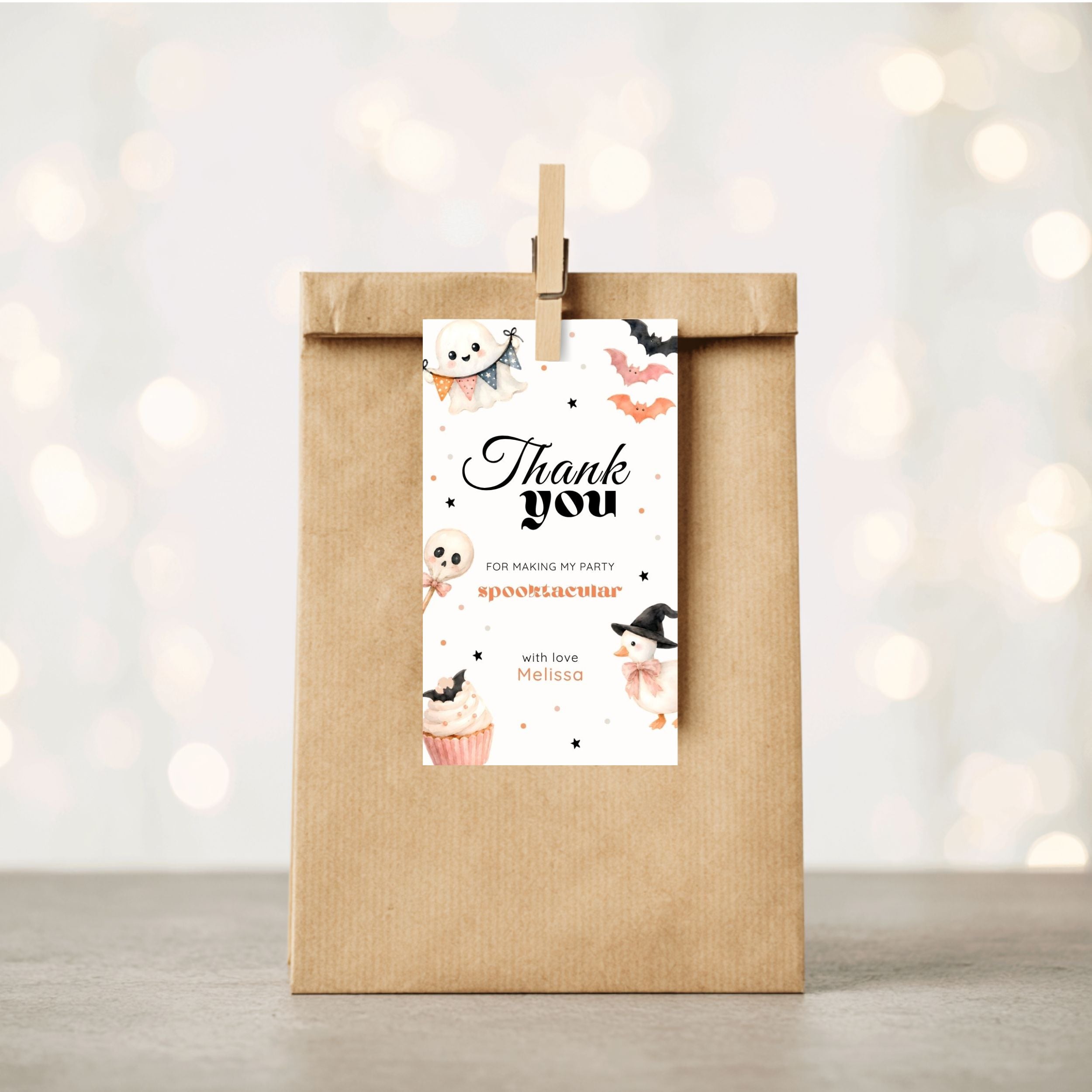 A kraft paper gift bag stands upright on a light surface with a wooden clip holding a printed thank-you tag. The tag has Halloween-themed illustrations and the text 'Thank you for making my party spooktacular' with the name 'Mia'.