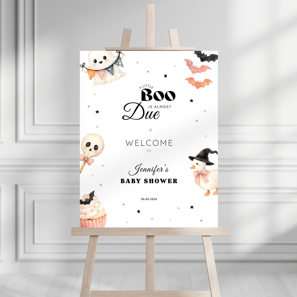 A Halloween-themed baby shower sign displayed on a wooden easel. The white sign features cute watercolor illustrations of a ghost, a cupcake, a chick in a witch hat, bats, and stars, with the text 'A Little Boo Is Almost Due Welcome to Jennifer's Baby Shower' and the date '06.