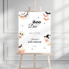 A Halloween-themed baby shower sign displayed on a wooden easel. The white sign features cute watercolor illustrations of a ghost, a cupcake, a chick in a witch hat, bats, and stars, with the text 'A Little Boo Is Almost Due Welcome to Jennifer's Baby Shower' and the date '06.