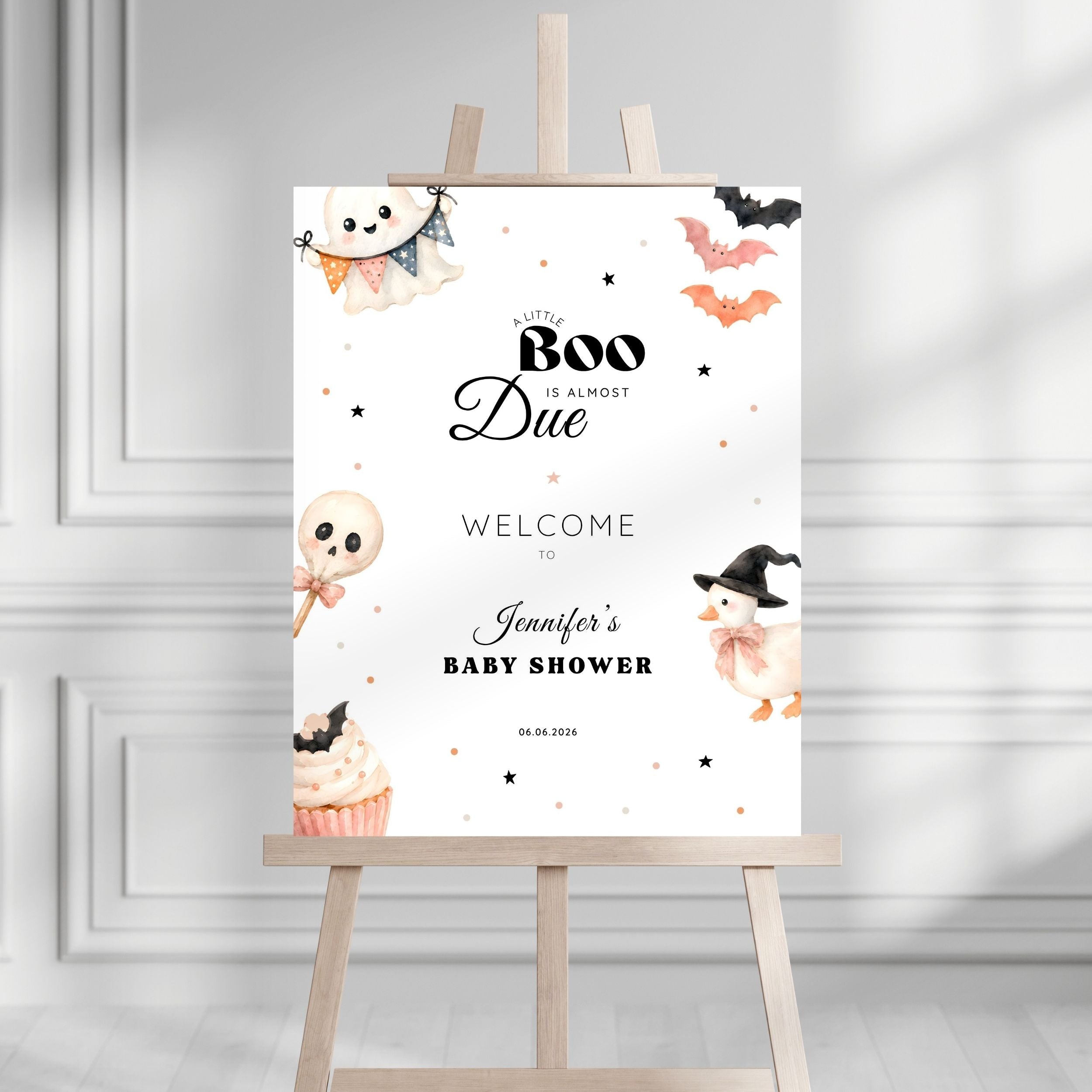A Halloween-themed baby shower sign displayed on a wooden easel. The white sign features cute watercolor illustrations of a ghost, a cupcake, a chick in a witch hat, bats, and stars, with the text 'A Little Boo Is Almost Due Welcome to Jennifer's Baby Shower' and the date '06.