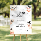 A Halloween-themed baby shower welcome sign on a wooden easel, featuring cute watercolor illustrations of a ghost, lollipop skull, cupcake, and a witch duck, set against a blurred outdoor garden background.