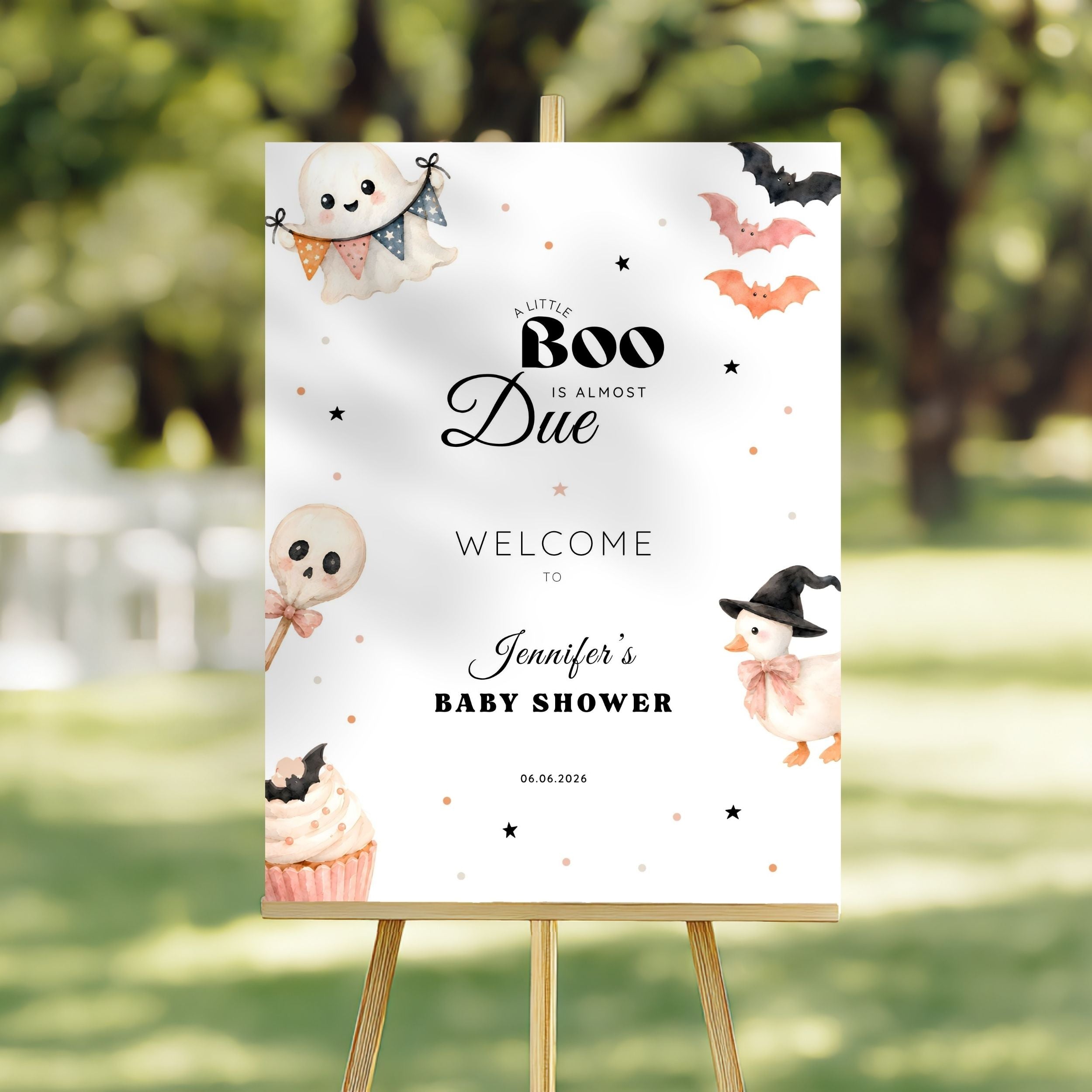 A Halloween-themed baby shower welcome sign on a wooden easel, featuring cute watercolor illustrations of a ghost, lollipop skull, cupcake, and a witch duck, set against a blurred outdoor garden background.