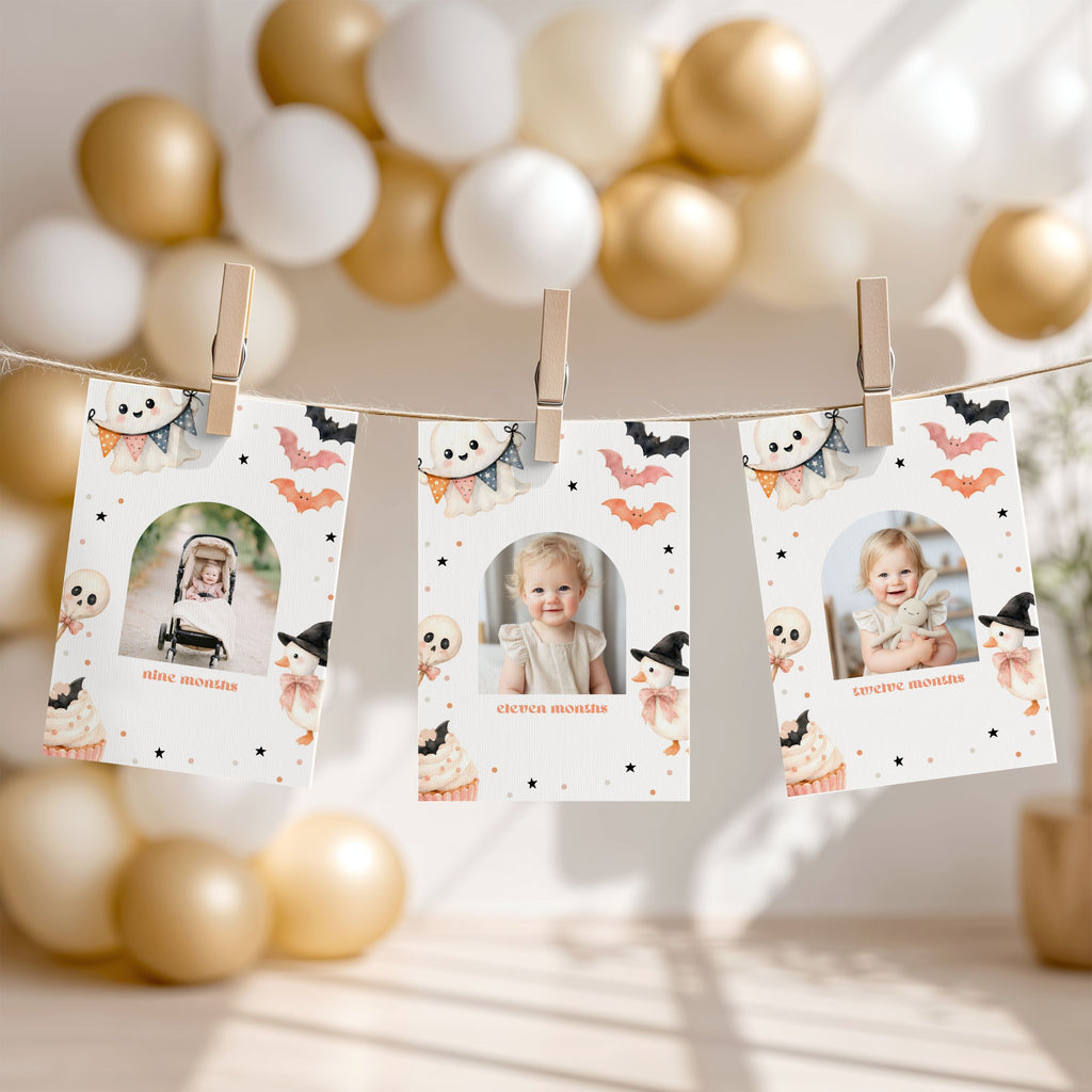 Three printed photo cards clipped to a string with wooden clothespins, decorated with Halloween motifs like bats, ghosts, and cupcakes. The cards hang in front of a softly blurred background of gold and white balloons.