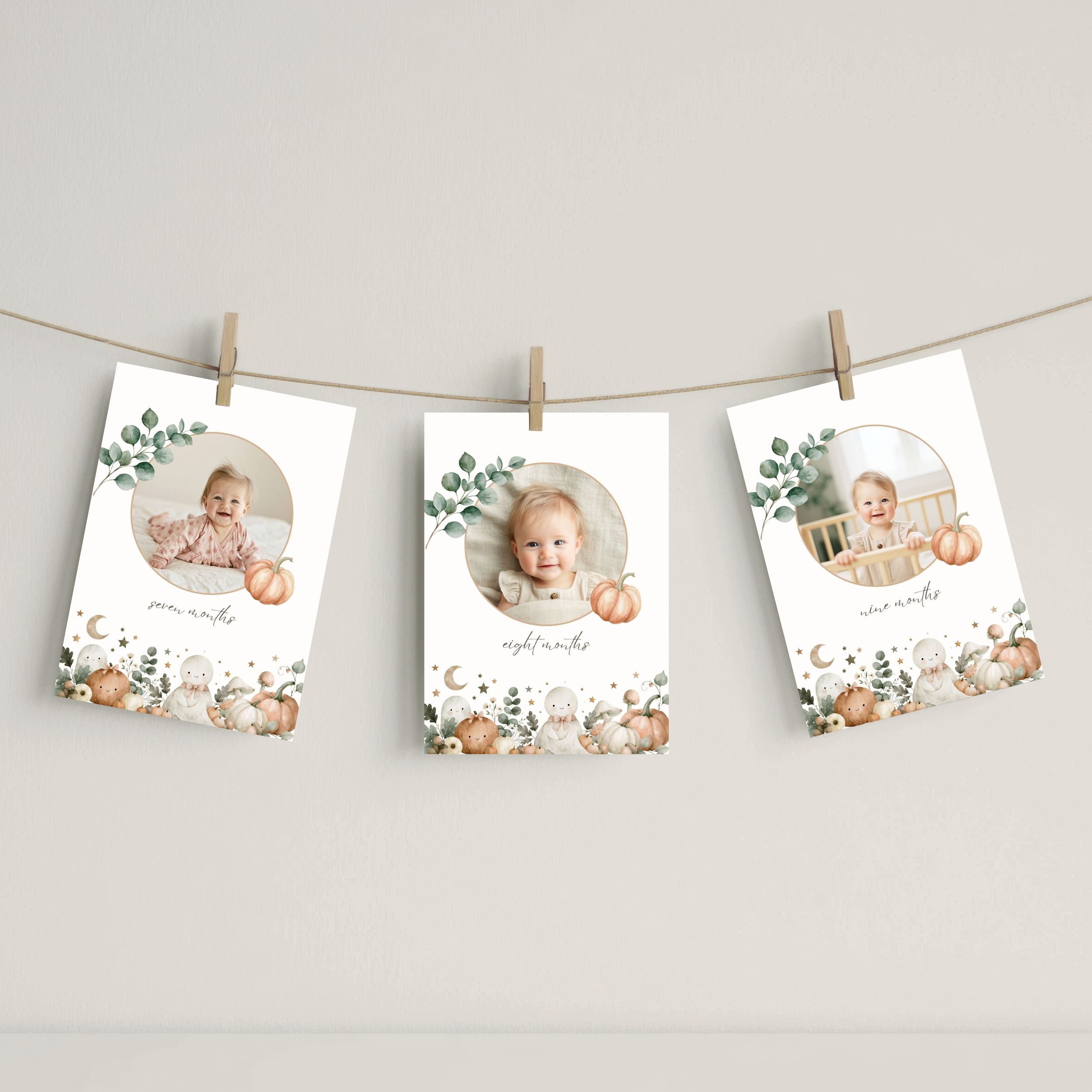 Three vertical halloween baby milestone photo cards with autumn watercolor designs are clipped to a string against a neutral wall. Each card features a circular photo of a baby, a pumpkin, eucalyptus leaves, and cute animal illustrations at the bottom.