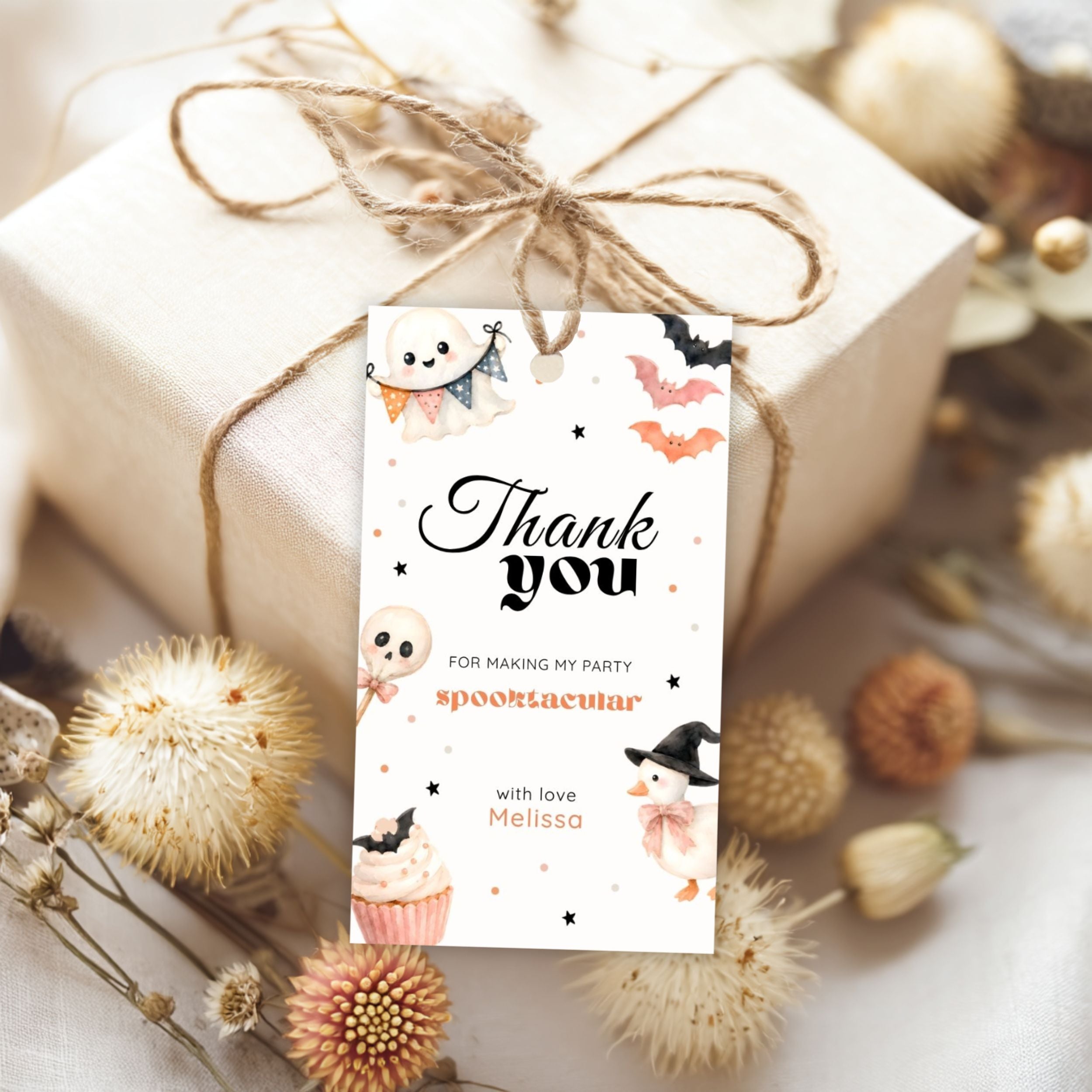 A wrapped gift box tied with twine and a hanging thank-you card with Halloween and spooky ghost illustrations, styled with dried flowers on a light fabric background.