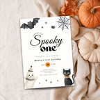 A Halloween-themed first birthday invitation card styled in a flat lay with autumn props. The white card features illustrations of a bat, spider web, spider, ghost, black cat, and stars, with the text 'Wesley is a Spooky one' and event details.