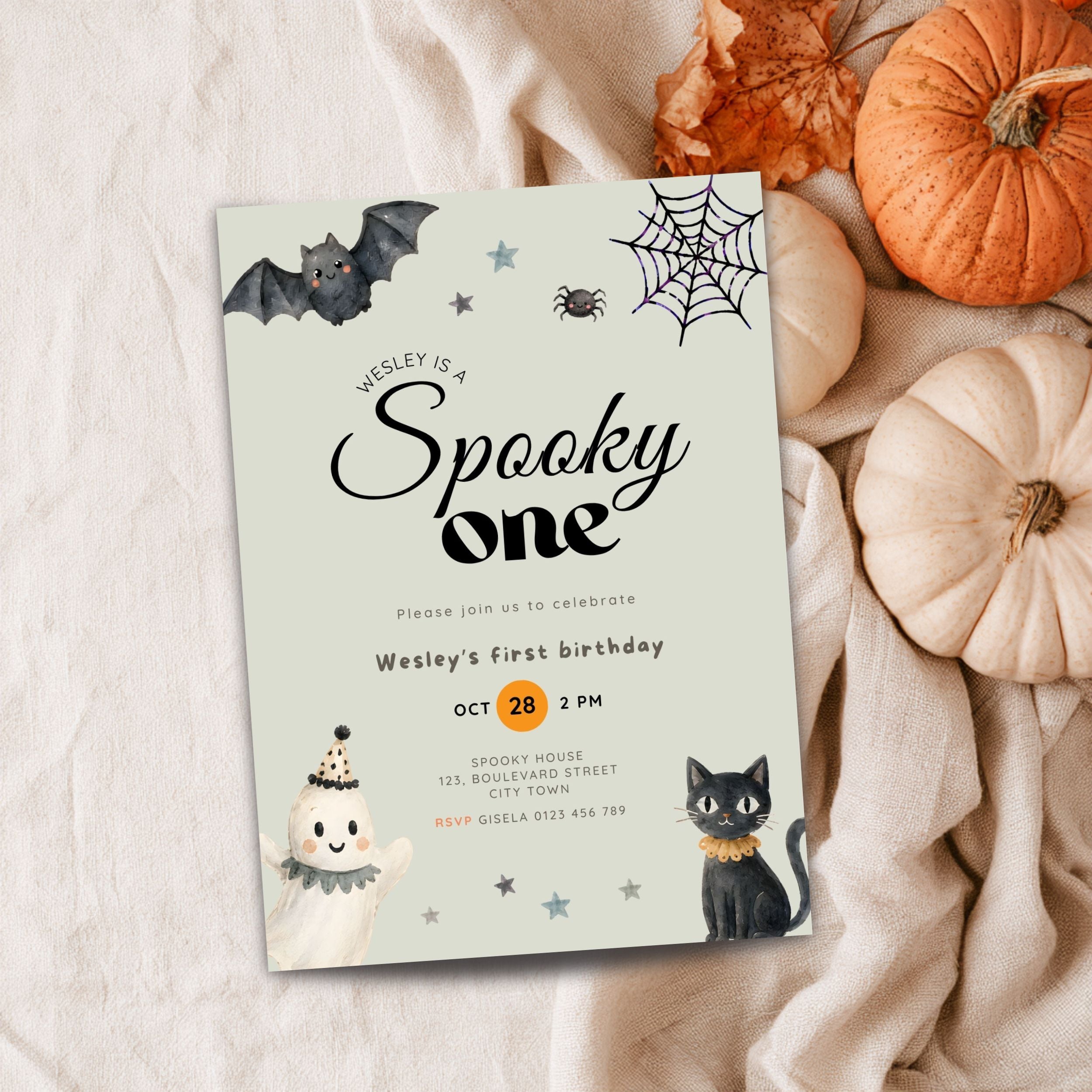A Halloween-themed first birthday invitation card styled in a flat lay with autumn props. The card features illustrations of a bat, spider, ghost, black cat, and stars on a pale sage green background.