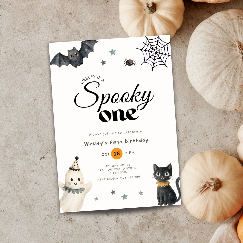A Halloween-themed first birthday invitation card styled on a textured gray surface with decorative pumpkins around it. The card features black script text reading 'Wesley is a Spooky one' with illustrations of a bat, spiderweb, spider, ghost, black cat, and stars.