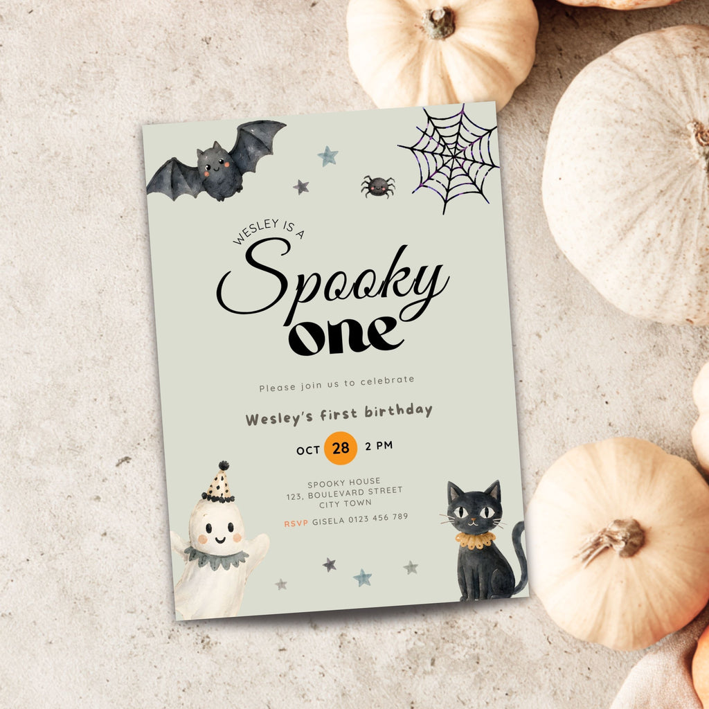 A Halloween-themed first birthday invitation card styled on a textured light gray surface with decorative pumpkins around it. The card features a pale mint background with black bat, spider, and moon illustrations, plus a black cat and ghost illustrations.