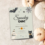 A Halloween-themed first birthday invitation card styled on a textured light gray surface with decorative pumpkins around it. The card features a pale mint background with black bat, spider, and moon illustrations, plus a black cat and ghost illustrations.