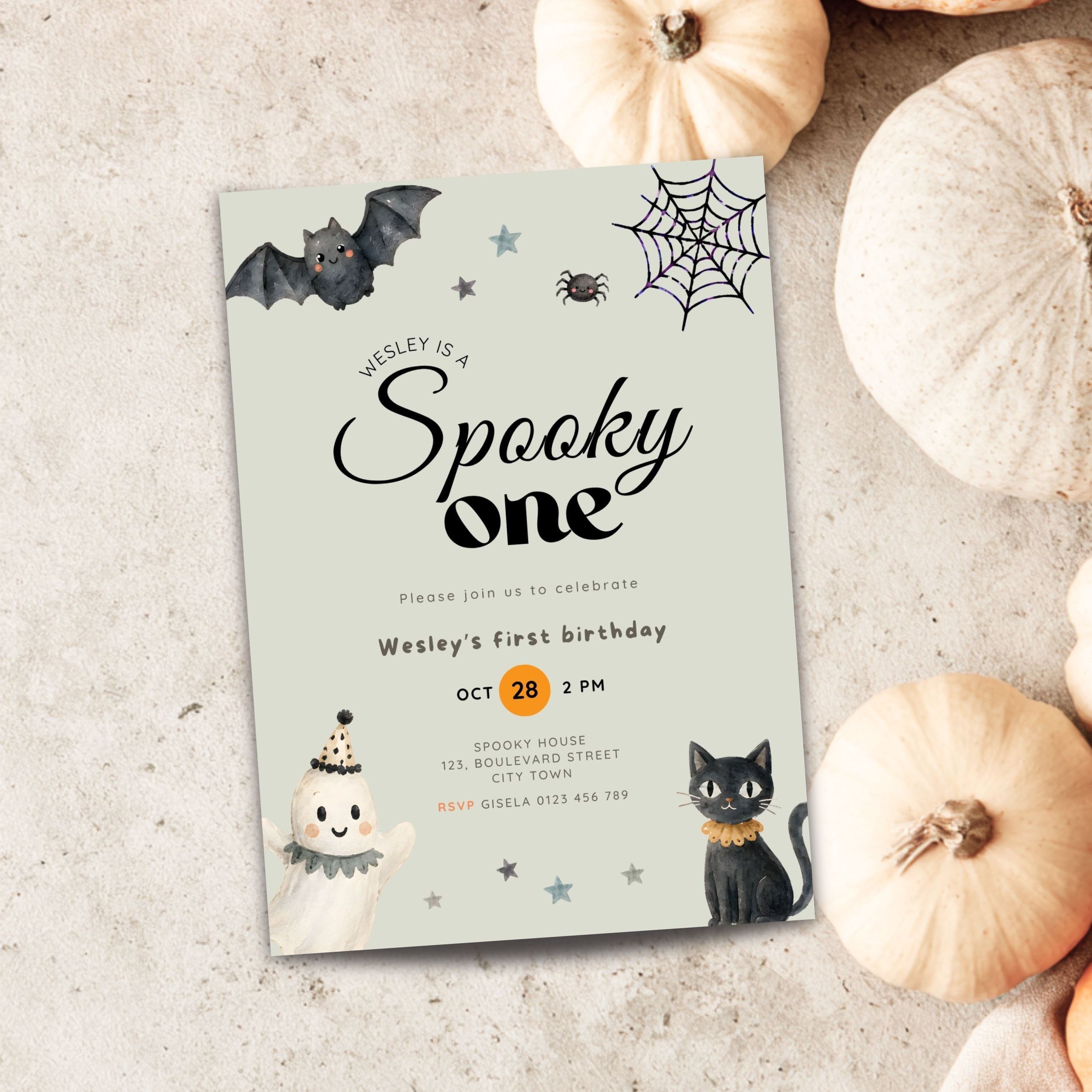 A Halloween-themed first birthday invitation card styled on a textured light gray surface with decorative pumpkins around it. The card features a pale mint background with black bat, spider, and moon illustrations, plus a black cat and ghost illustrations.