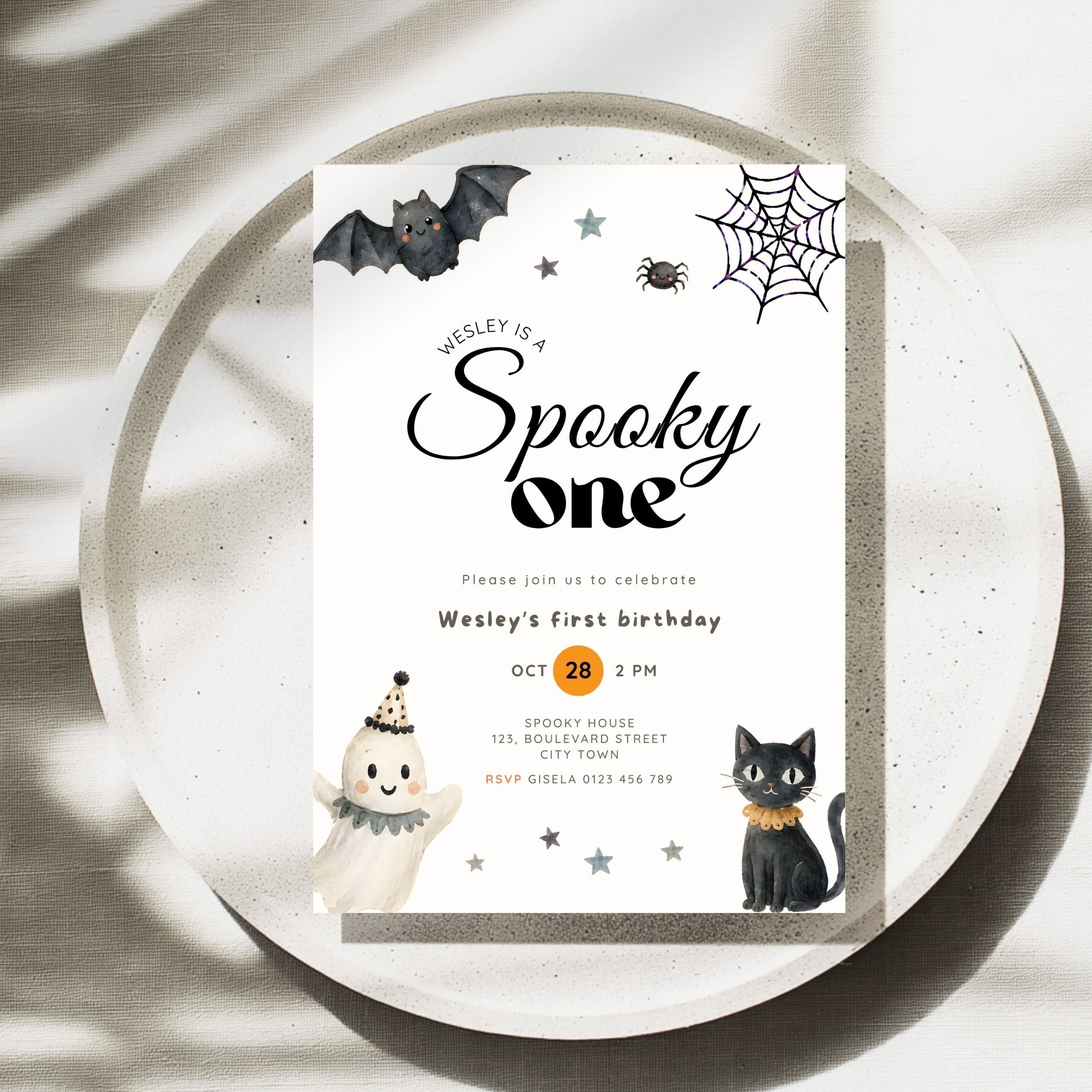 A square Halloween-themed birthday invitation card for Wesley's first birthday, styled flat-lay on a round textured plate with soft leaf shadows. The card features cute illustrations of a bat, spider, spiderweb, ghost, black cat, and stars in black, white, orange, and gray.