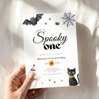 A hand holds a Halloween-themed first birthday invitation card featuring a bat, spider, moon, ghost, and black cat illustrations on a white fabric background.