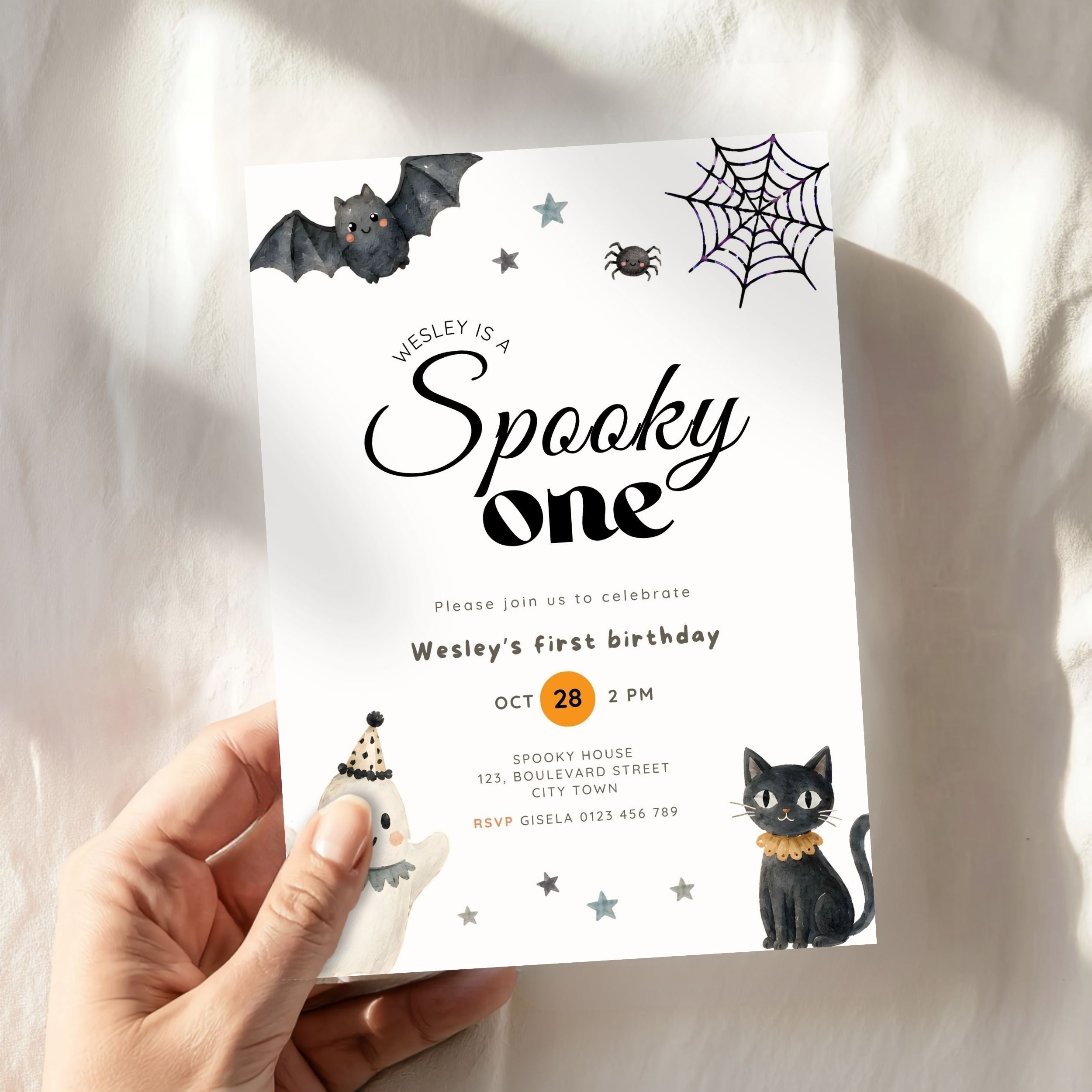 A hand holds a Halloween-themed first birthday invitation card featuring a bat, spider, moon, ghost, and black cat illustrations on a white fabric background.