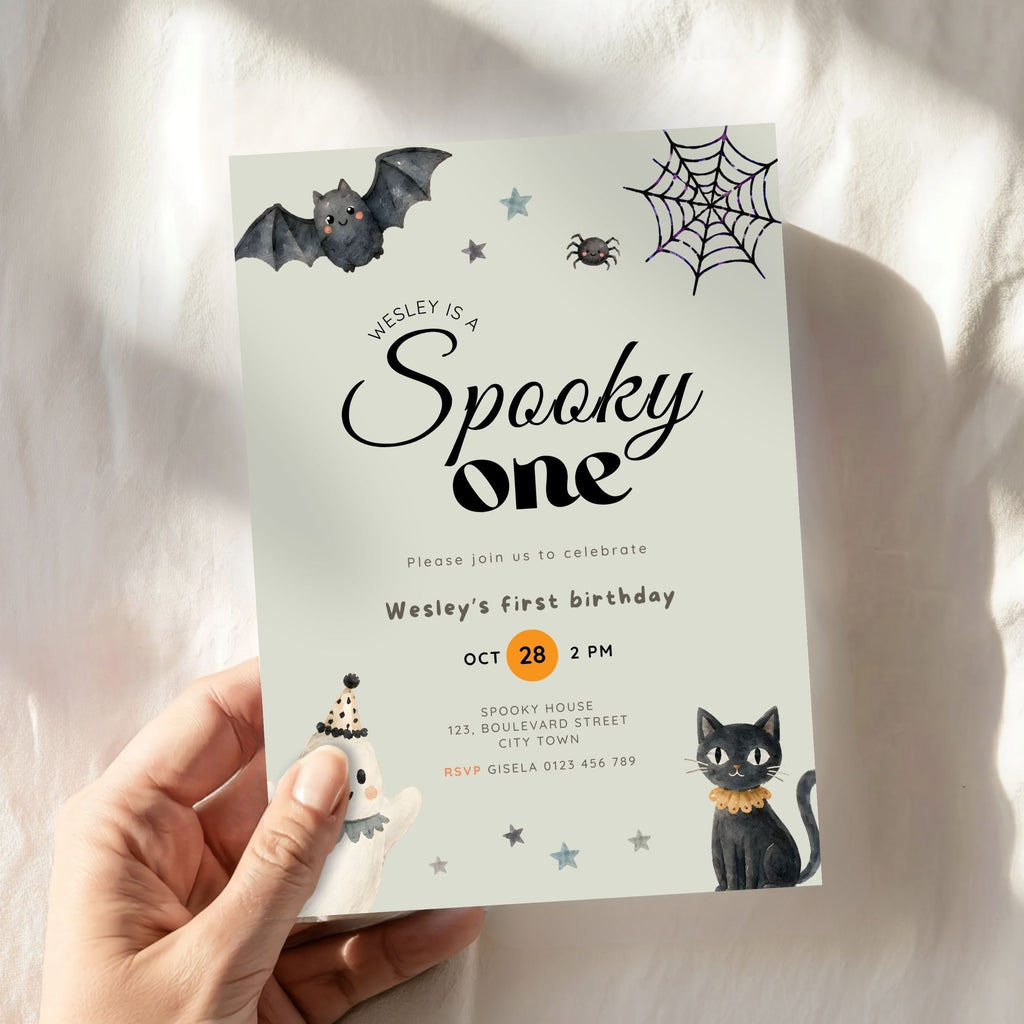 A hand holds a Halloween-themed first birthday invitation card against a softly draped white fabric background. The card features a pale mint background with black text reading 'Wesley is a Spooky one,' decorated with illustrations of a bat, spiderweb, spider, cat, and a small ghost.
