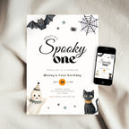 A Halloween-themed birthday invitation card for Wesley's first birthday, styled on a beige fabric background next to a smartphone displaying the digital version of the invitation. The card features black text, illustrations of a bat, spiderweb, spider, ghost, and black cat, with a small orange circle highlighting the date.