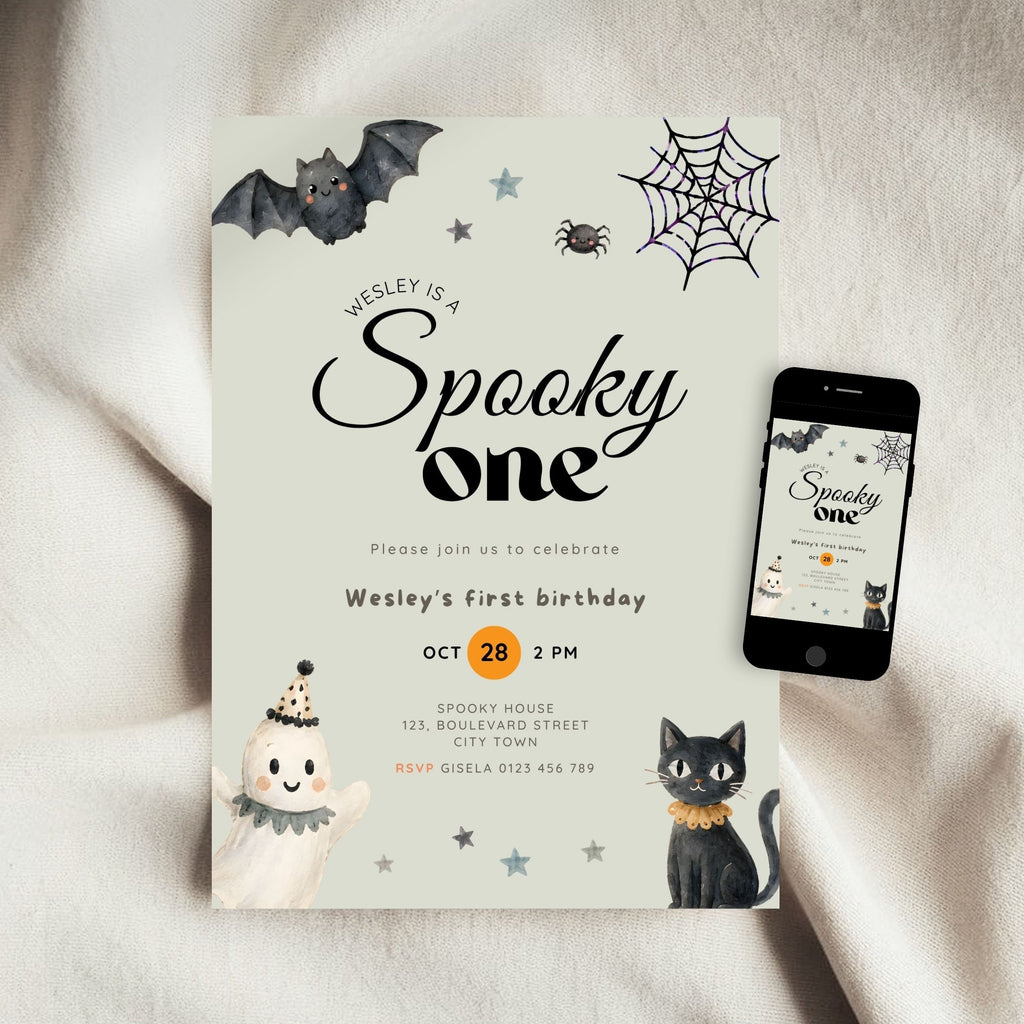 A Halloween-themed first birthday invitation card displayed on a soft beige fabric background, next to a smartphone showing the same design on a screen. The card features spooky illustrations including a bat, spiderweb, spider, cat, and ghost.