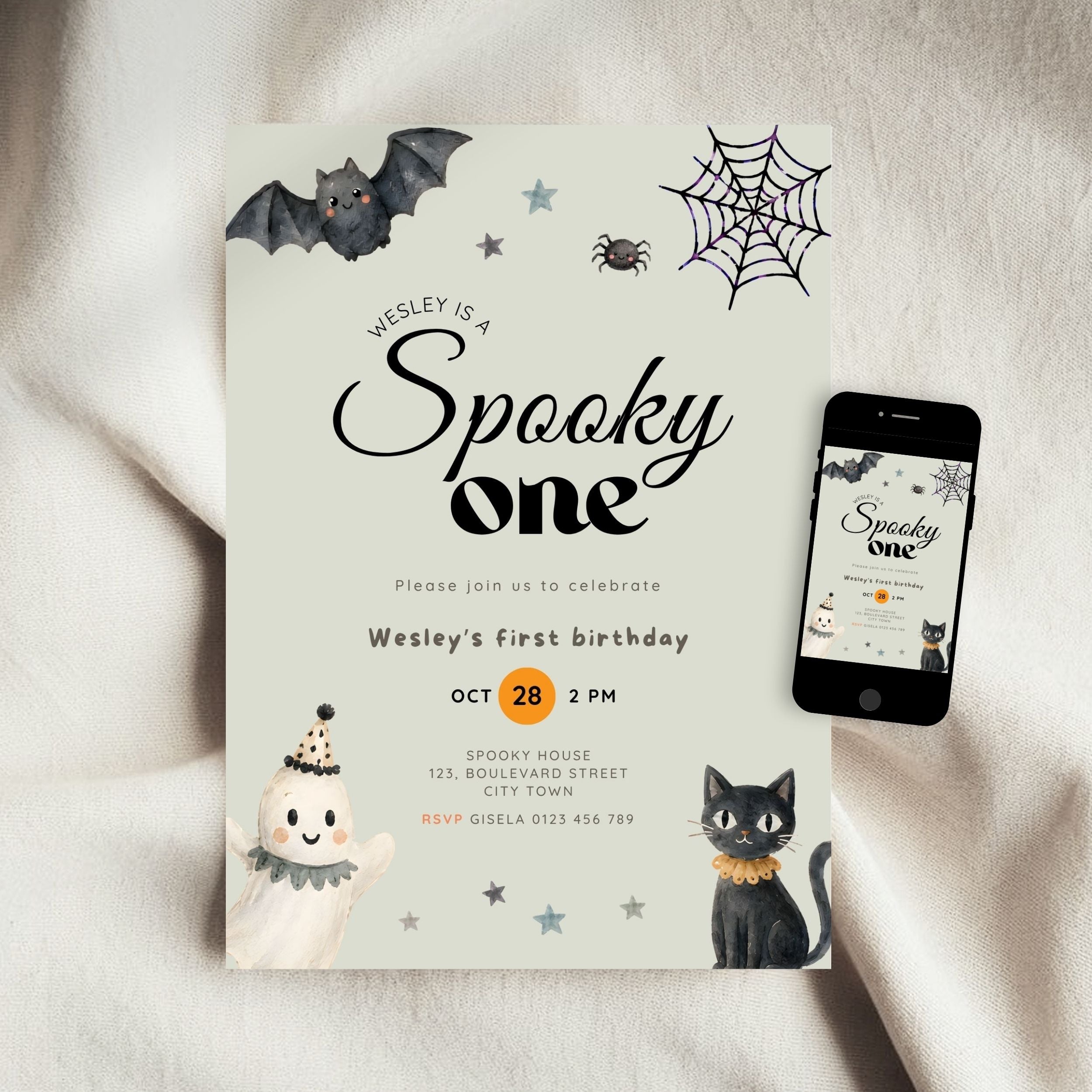 A Halloween-themed first birthday invitation card displayed on a soft beige fabric background, next to a smartphone showing the same design on a screen. The card features spooky illustrations including a bat, spiderweb, spider, cat, and ghost.