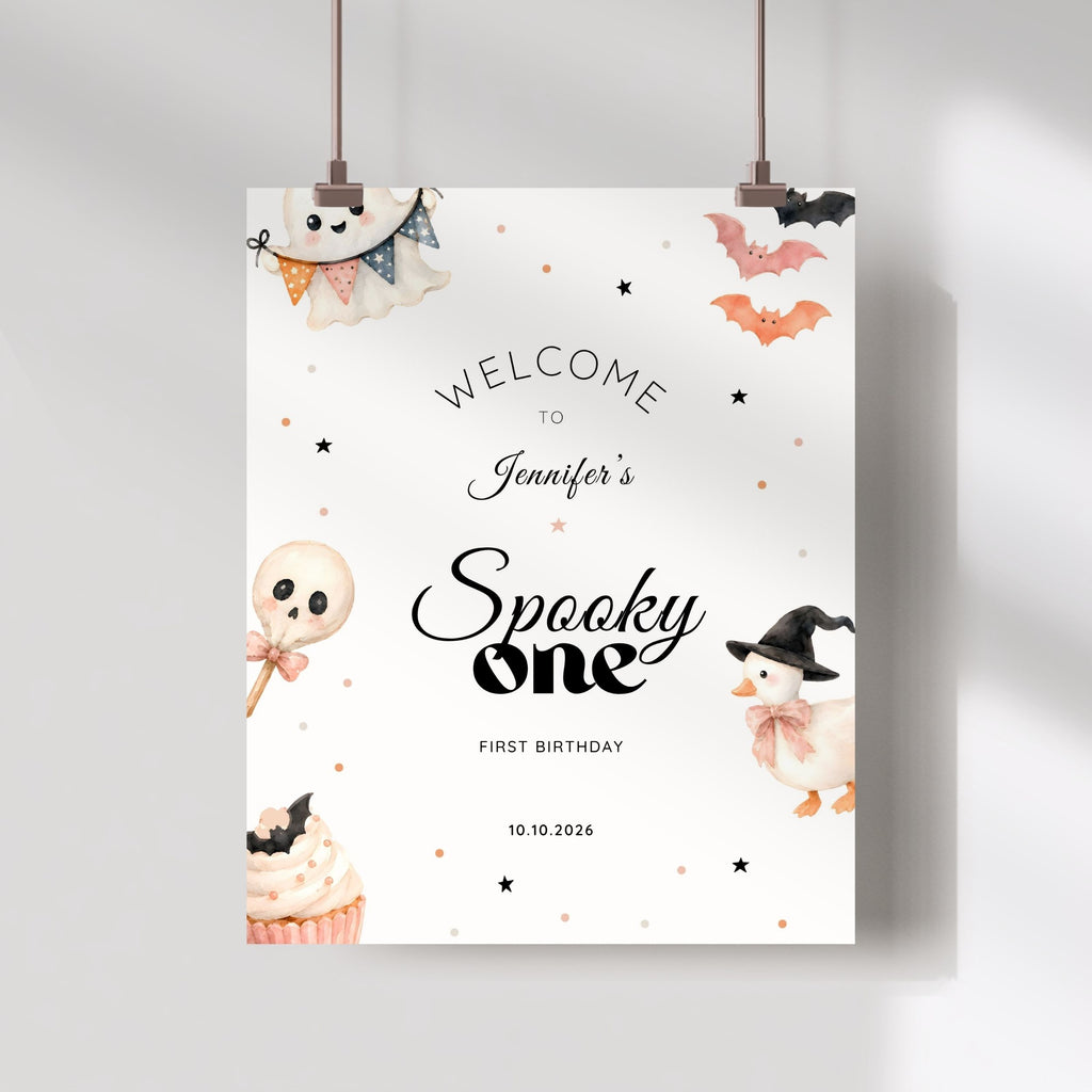 A personalized Halloween party sign hanging from two clips against a light gray wall. The sign has a white background with cute watercolor illustrations of a ghost, cupcake, lollipop, duck, and bats, along with the text 'WELCOME TO Jennifer's Spooky one FIRST BIRTHDAY' and the date '10.