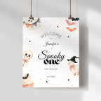 A personalized Halloween party sign hanging from two clips against a light gray wall. The sign has a white background with cute watercolor illustrations of a ghost, cupcake, lollipop, duck, and bats, along with the text 'WELCOME TO Jennifer's Spooky one FIRST BIRTHDAY' and the date '10.