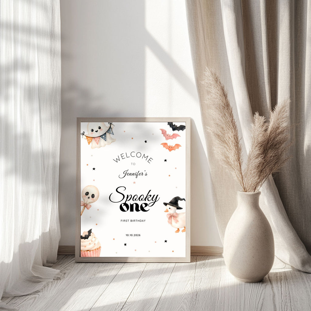 A framed Halloween-themed welcome sign for a 'Spooky One' birthday stands on a light wood floor between sheer white curtains and a beige ceramic vase with dried pampas grass. The sign features cute illustrations of a ghost, skull, witch, cupcake, and bats on a white background.
