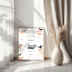 A framed Halloween-themed welcome sign for a 'Spooky One' birthday stands on a light wood floor between sheer white curtains and a beige ceramic vase with dried pampas grass. The sign features cute illustrations of a ghost, skull, witch, cupcake, and bats on a white background.