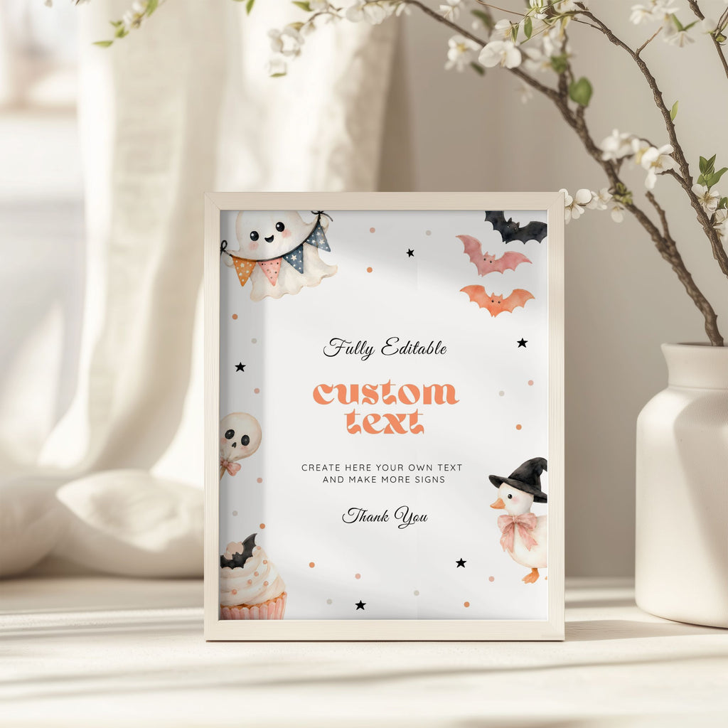 A framed Halloween-themed editable sign mockup standing on a tabletop. The white sign features cute watercolor illustrations of a ghost, pumpkin, bat, snowman, and cupcake, with placeholder text reading 'custom text' and other decorative text.