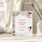 A framed Halloween-themed editable sign mockup standing on a tabletop. The white sign features cute watercolor illustrations of a ghost, pumpkin, bat, snowman, and cupcake, with placeholder text reading 'custom text' and other decorative text.