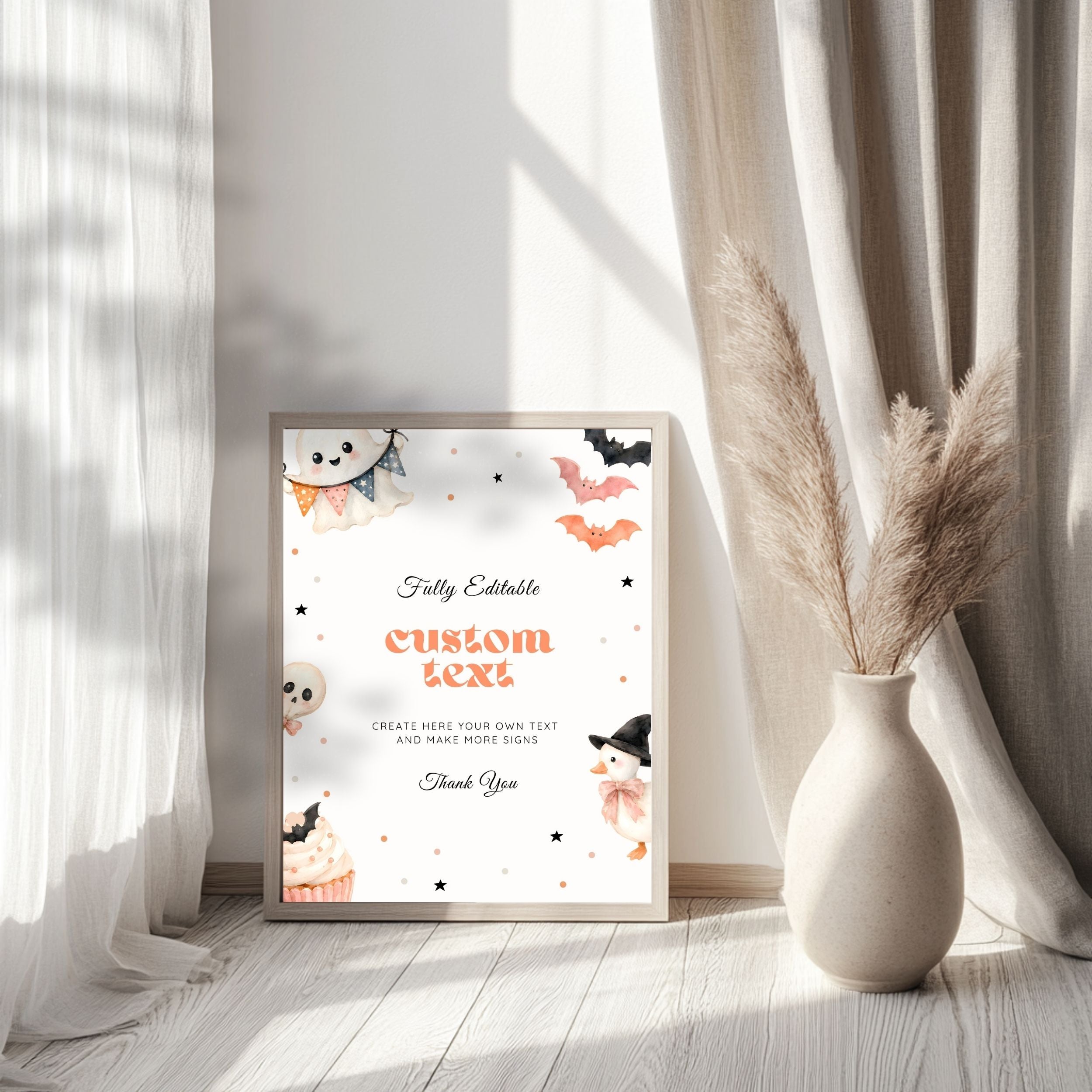 A framed Halloween-themed custom print leaning against a wall on a light wood floor, styled in a room with sheer white curtains, beige drapery, and a ceramic vase with pampas grass. The print features watercolor illustrations of a ghost, bats, a cupcake, and a witch hat on a white background with orange and black text.