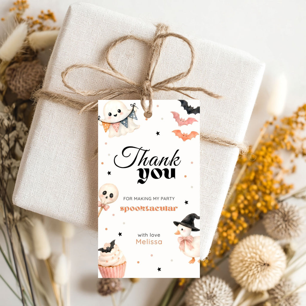 A wrapped gift box tied with twine and adorned with a Halloween-themed thank you tag featuring cute ghost, bat, and pumpkin illustrations. The tag reads 'Thank you FOR MAKING MY PARTY spooktacular with love Melissa'.