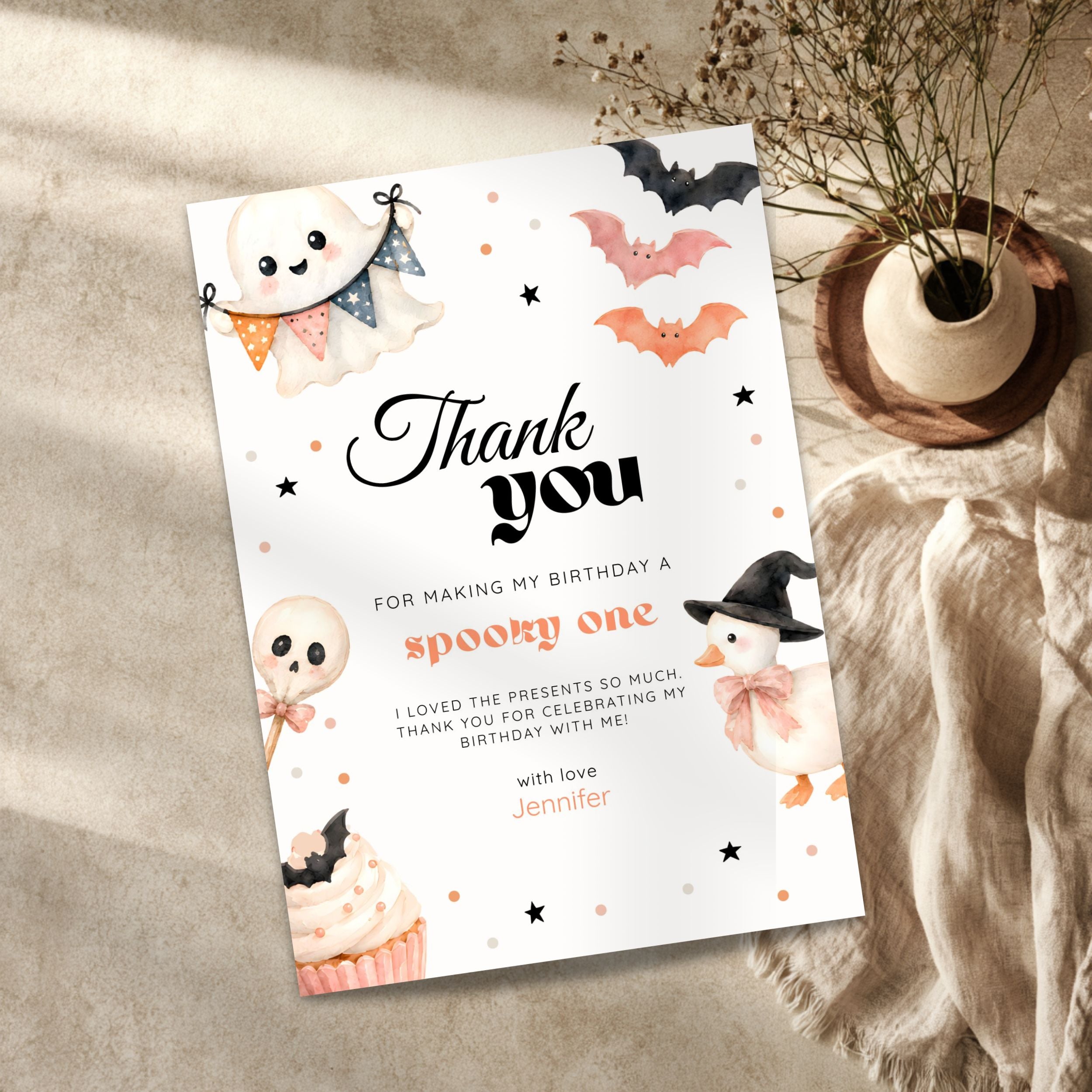 A Halloween-themed birthday thank you card styled in a flat-lay setting on a textured beige surface, surrounded by a small potted plant and a beige fabric runner.