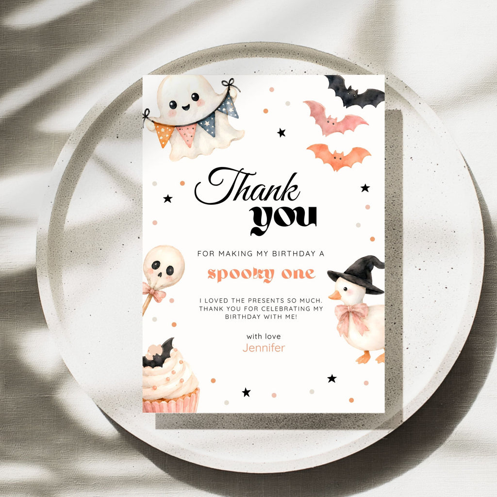 A Halloween-themed thank you card with cute illustrations of a ghost, bats, a witch, a cupcake, and stars, placed on a round tray with soft shadow overlays.