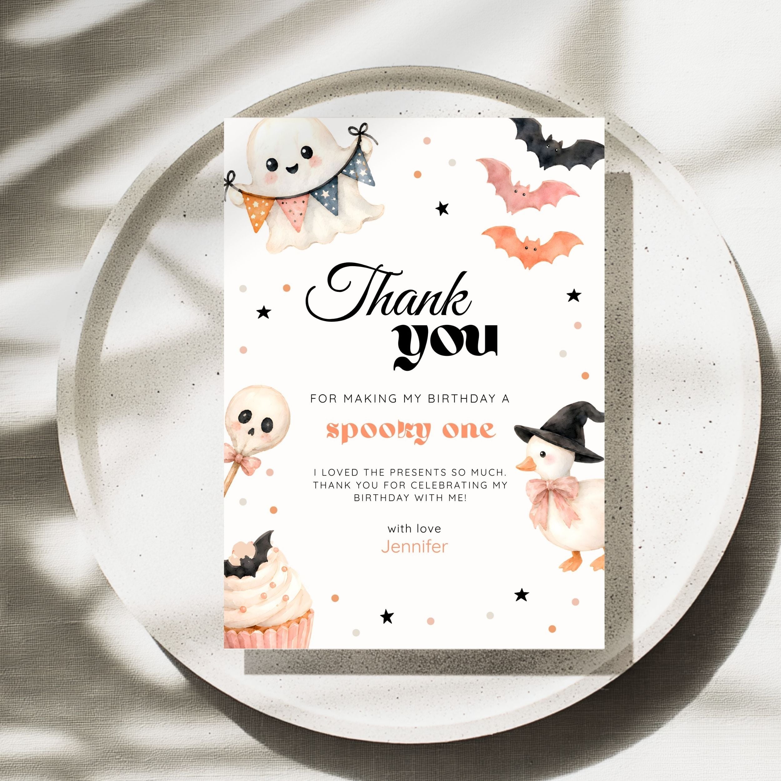 A Halloween-themed thank you card with cute illustrations of a ghost, bats, a witch, a cupcake, and stars, placed on a round tray with soft shadow overlays.
