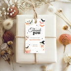 A wrapped gift box tied with twine and topped with a decorative 'Thank you' hang tag featuring Halloween illustrations, photographed in a styled flat-lay with dried flowers and autumn botanicals.