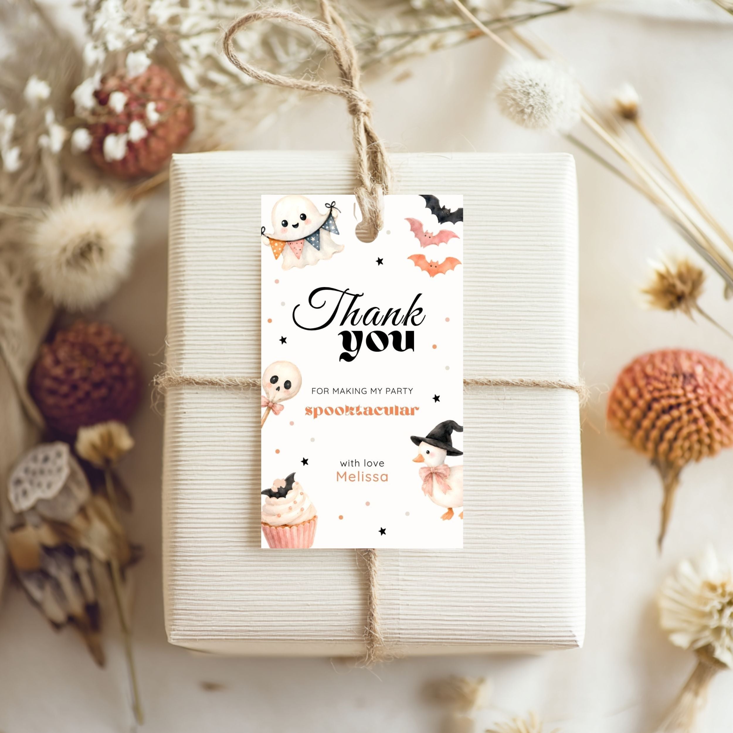 A wrapped gift box tied with twine and topped with a decorative 'Thank you' hang tag featuring Halloween illustrations, photographed in a styled flat-lay with dried flowers and autumn botanicals.