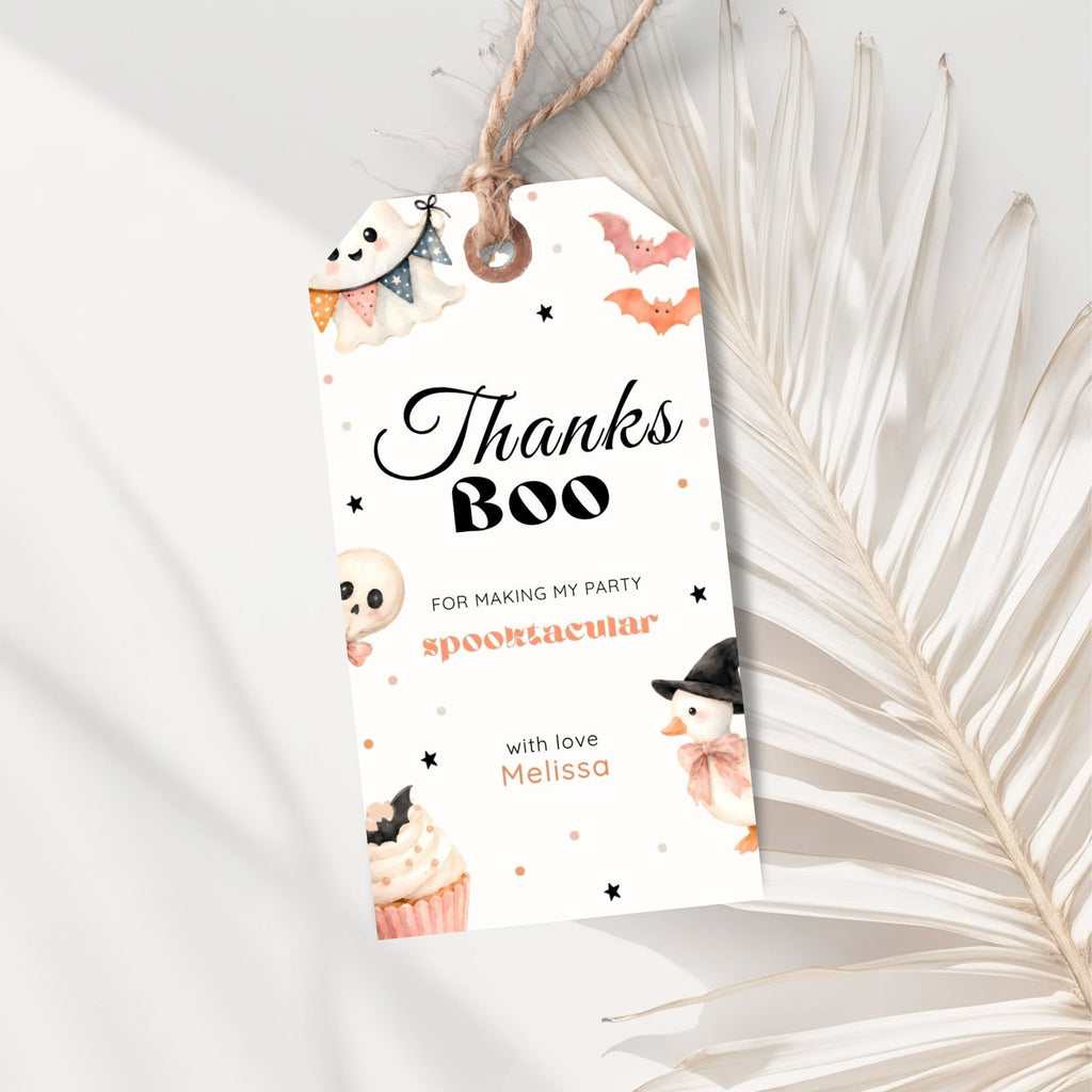 A Halloween-themed gift tag with a rustic twine loop, featuring cute illustrations of a ghost, bat, cupcake, and snowman, with the text 'Thanks Boo' on a white background styled with dried palm leaves.