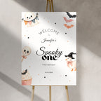 A personalized Halloween welcome sign displayed on a wooden easel against a textured gray wall. The sign features cute watercolor illustrations of a ghost, a witch, a pumpkin, a lollipop skull, a cupcake, and bats, with the text 'WELCOME TO Jennifer's Spooky one FIRST BIRTHDAY' and the date '10.