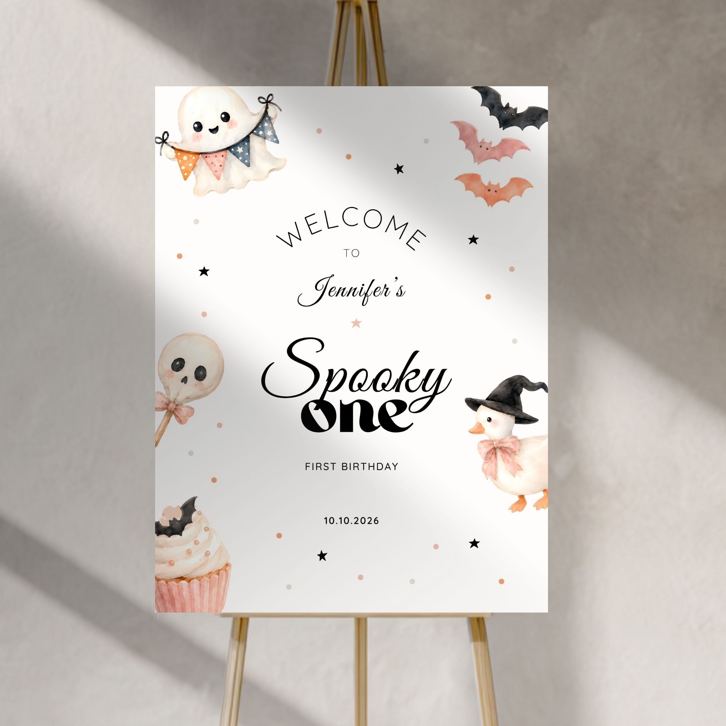 A personalized Halloween welcome sign displayed on a wooden easel against a textured gray wall. The sign features cute watercolor illustrations of a ghost, a witch, a pumpkin, a lollipop skull, a cupcake, and bats, with the text 'WELCOME TO Jennifer's Spooky one FIRST BIRTHDAY' and the date '10.