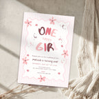 A pink and white 'One Happy Girl' one-year birthday invitation card styled on a textured beige surface with a cream knit blanket and dried grass.