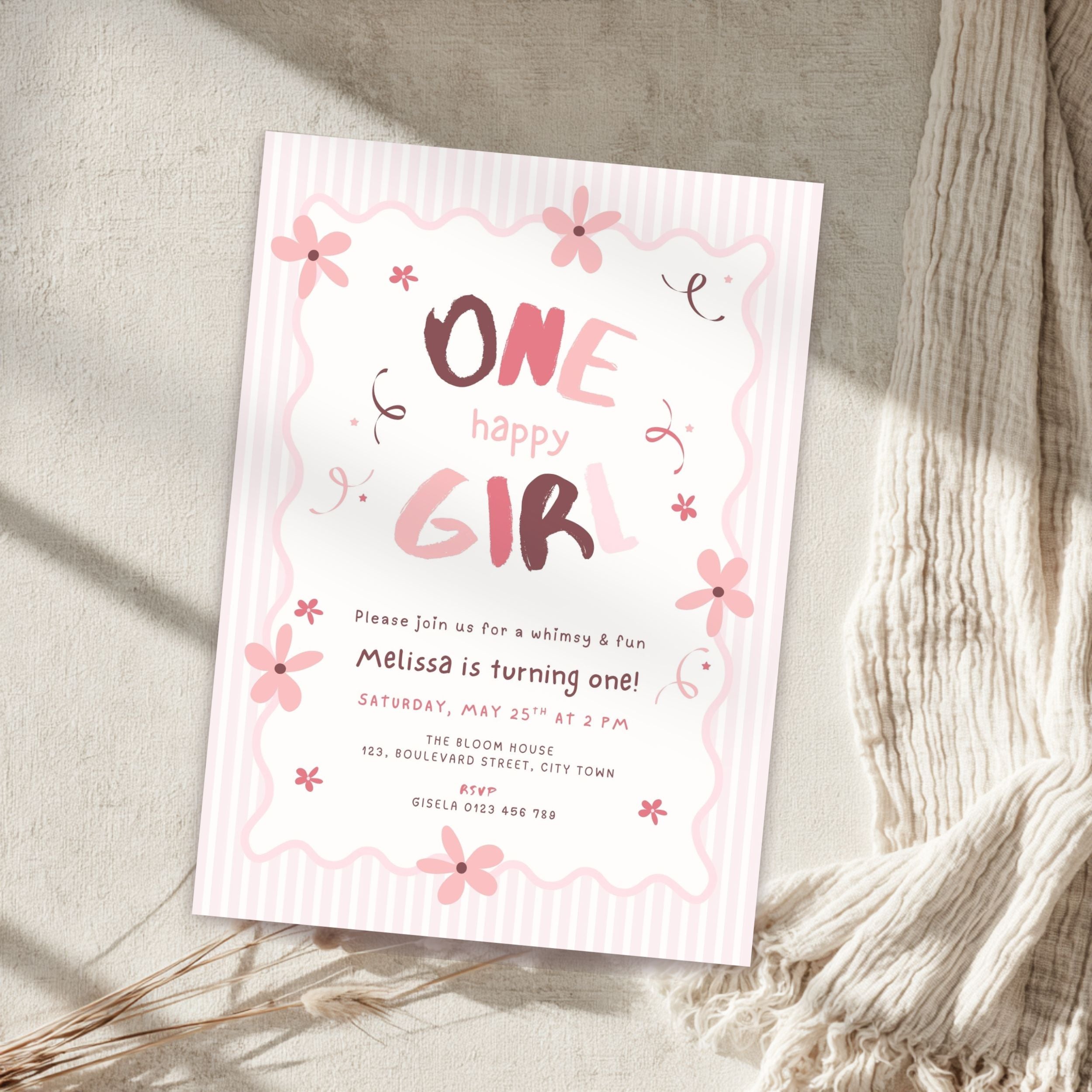 A pink and white 'One Happy Girl' one-year birthday invitation card styled on a textured beige surface with a cream knit blanket and dried grass.