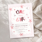 A pink and white striped 'One Happy Girl' 1st birthday invitation card with floral illustrations, styled on a textured beige fabric background.