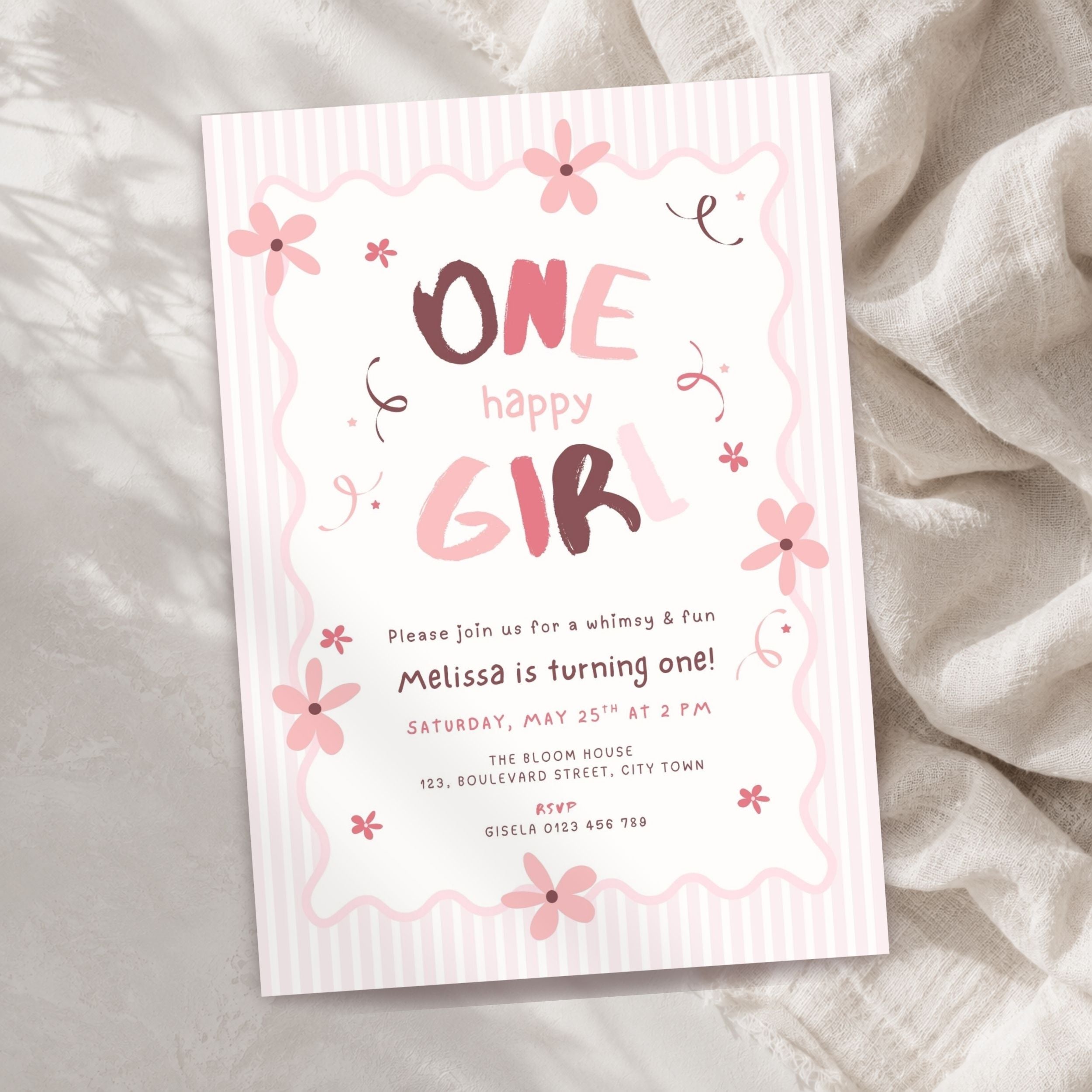 A pink and white striped 'One Happy Girl' 1st birthday invitation card with floral illustrations, styled on a textured beige fabric background.